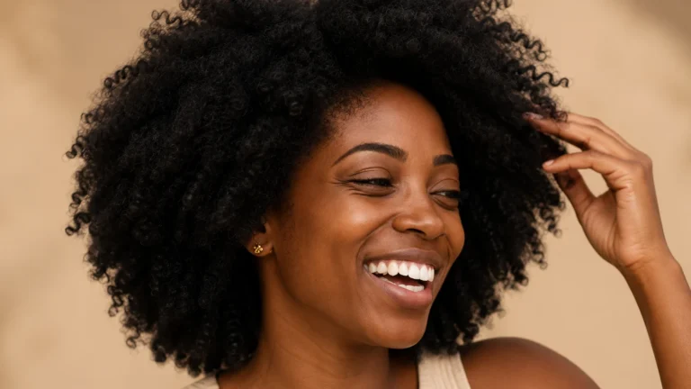 Mulher negra sorrindo e tocando o cabelo crespo natural em retrato com fundo bege.